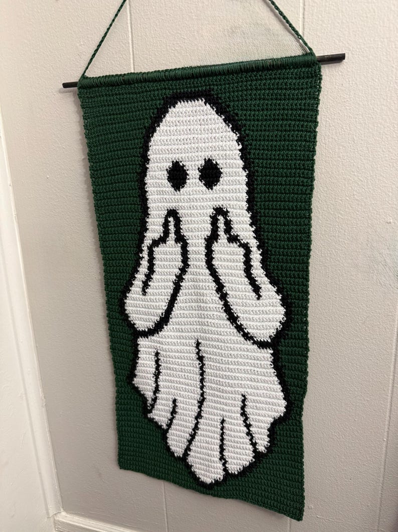 May include: A rectangular crochet wall hanging featuring a white ghost figure on a dark green background. The ghost has black eyes and is depicted with both arms raised, showing middle fingers. The piece is suspended from a thin black rod.