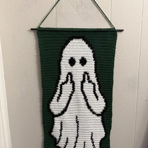 May include: A rectangular crochet wall hanging featuring a white ghost figure centered on a dark green background. The ghost has two black circular eyes and is depicted with both arms raised, showing a middle finger gesture on each hand.