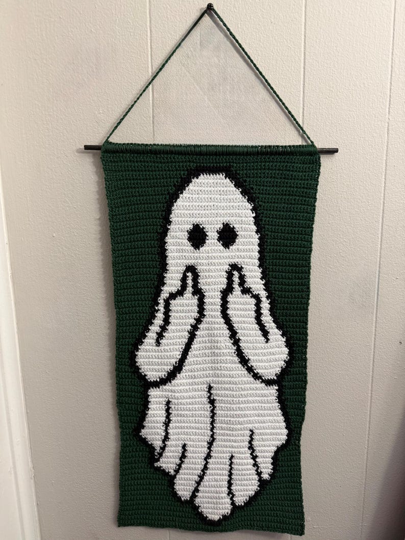 May include: A rectangular crochet wall hanging featuring a white ghost figure centered on a dark green background. The ghost has two black circular eyes and is depicted with both arms raised, showing a middle finger gesture on each hand.