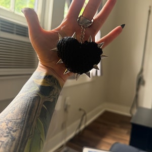 May include: A black crocheted heart pendant with silver spikes around the edges. The heart is suspended from a silver chain and ring. The pendant is held up in front of a window.