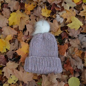 Puede incluir: Un gorro de punto morado con un pompón blanco y esponjoso en la parte superior, que se encuentra sobre una cama de hojas de otoño caídas.
