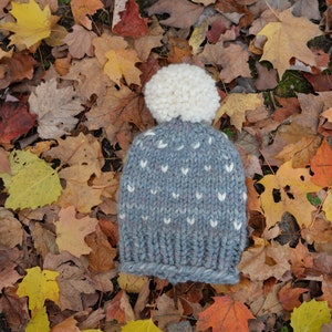 Puede incluir: Un gorro de punto gris con un pompón blanco y un patrón de corazones blancos. El gorro está sobre un lecho de hojas de otoño caídas.