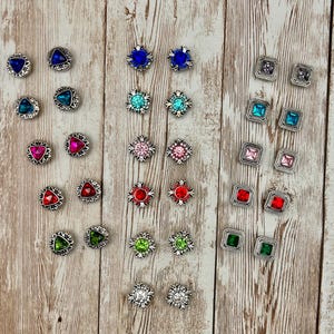 May include: Assortment of colorful rhinestone embellishments in various shapes and sizes. The embellishments are silver-toned and feature a variety of colors, including blue, green, red, pink, and clear.