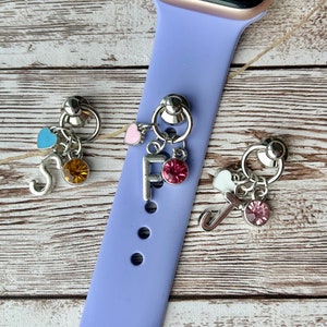 May include: A purple Apple Watch band with three charms attached. The charms are a silver letter 'S', a silver letter 'F', and a silver umbrella with a pink crystal.