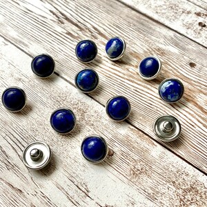 K&ouml;nnte beinhalten: Eine Ansammlung runder, dunkelblauer Lapis-Lazuli-Edelstein-Druckkn&ouml;pfe mit silberfarbenen Metallr&auml;ndern und -r&uuml;ckseiten. Die Druckkn&ouml;pfe sind auf einer hellen, verwitterten Holzoberfl&auml;che verteilt, was eine nat&uuml;rliche und elegante &Auml;sthetik erzeugt.