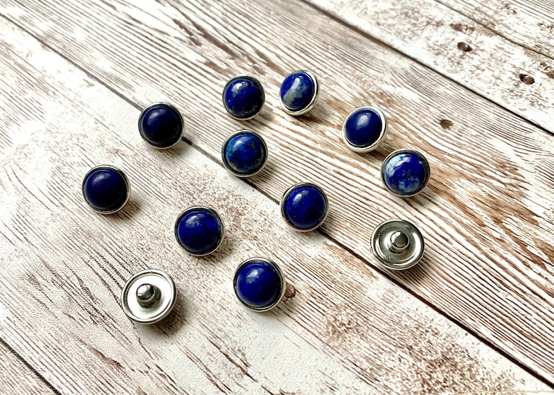 K&ouml;nnte beinhalten: Eine Ansammlung runder, dunkelblauer Lapis-Lazuli-Edelstein-Druckkn&ouml;pfe mit silberfarbenen Metallr&auml;ndern und -r&uuml;ckseiten. Die Druckkn&ouml;pfe sind auf einer hellen, verwitterten Holzoberfl&auml;che verteilt, was eine nat&uuml;rliche und elegante &Auml;sthetik erzeugt.