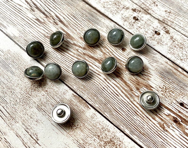 K&ouml;nnte beinhalten: Eine Sammlung runder, grau-gr&uuml;ner Edelstein-Druckkn&ouml;pfe mit silberfarbenen Metallr&auml;ndern und -r&uuml;ckseiten. Die Kn&ouml;pfe sind auf einer hellbraunen Holzoberfl&auml;che verstreut und zeigen ihr poliertes Aussehen.