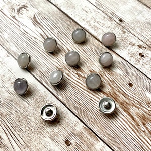 K&ouml;nnte beinhalten: Eine Sammlung runder Druckkn&ouml;pfe aus hellfarbigen Edelsteinen mit silberfarbenen Metallr&auml;ndern. Die Edelsteine variieren in den Farbt&ouml;nen Rosa, Lavendel und Grau. Die Druckkn&ouml;pfe sind auf einer hellbraunen Holzoberfl&auml;che angeordnet.