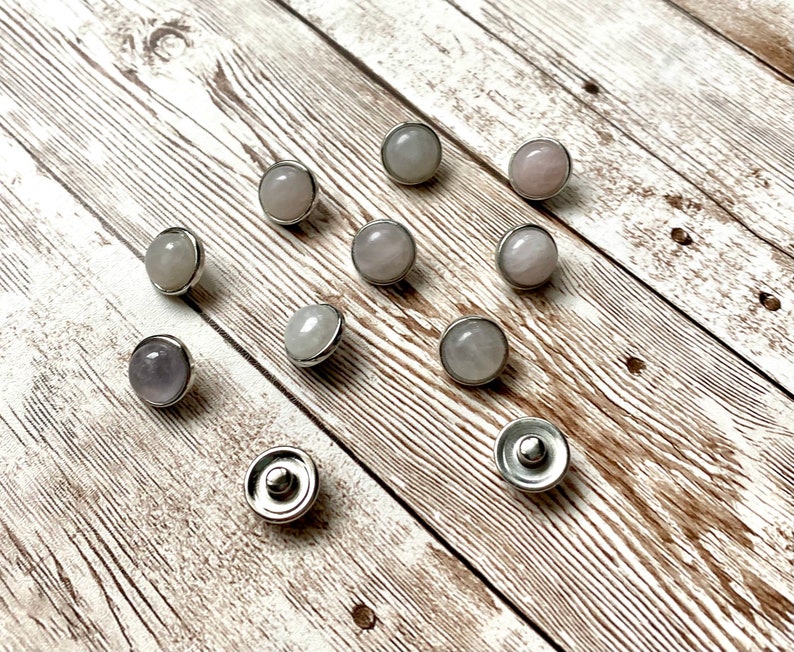 K&ouml;nnte beinhalten: Eine Sammlung runder Druckkn&ouml;pfe aus hellfarbigen Edelsteinen mit silberfarbenen Metallr&auml;ndern. Die Edelsteine variieren in den Farbt&ouml;nen Rosa, Lavendel und Grau. Die Druckkn&ouml;pfe sind auf einer hellbraunen Holzoberfl&auml;che angeordnet.
