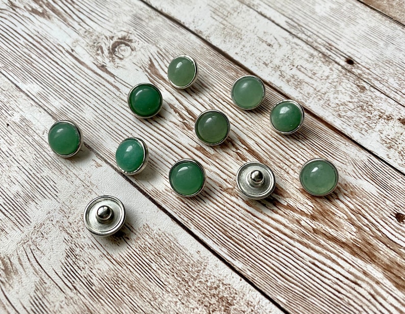 K&ouml;nnte beinhalten: Zehn gr&uuml;ne Edelstein-Druckkn&ouml;pfe mit silberfarbenen Metallr&auml;ndern und -r&uuml;ckseiten. Die runden Kn&ouml;pfe haben eine glatte, polierte Oberfl&auml;che. Sie sind auf einer hellen, strukturierten Holzoberfl&auml;che angeordnet.