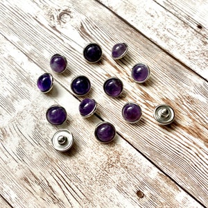 K&ouml;nnte beinhalten: Eine Ansammlung von Amethyst-Druckkn&ouml;pfen mit silberfarbenen Metallr&auml;ndern. Die violetten Edelsteine variieren in der Schattierung von hell bis tiefviolett. Die Kn&ouml;pfe sind auf einer hellen Holzmaserung verstreut.