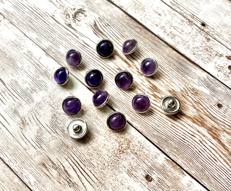 K&ouml;nnte beinhalten: Eine Ansammlung von Amethyst-Druckkn&ouml;pfen mit silberfarbenen Metallr&auml;ndern. Die violetten Edelsteine variieren in der Schattierung von hell bis tiefviolett. Die Kn&ouml;pfe sind auf einer hellen Holzmaserung verstreut.