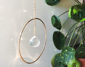 May include: A gold-toned metal suncatcher featuring a clear crystal ball within a circular frame, suspended by a chain. A houseplant with large, green leaves is visible in the background, next to the suncatcher.