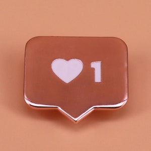 May include: Rose gold colored metal pin in the shape of a social media notification icon with a white heart and the number "1" inside.