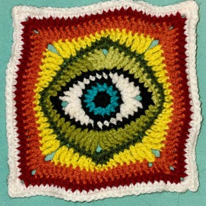 May include: A colorful crochet square with a central eye design. The eye is made of white, black, turquoise, and dark blue yarn. The square is surrounded by a border of red, orange, yellow, and green yarn.