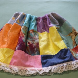 May include: A colourful patchwork skirt with a white lace trim. The skirt is made of different fabrics in shades of orange, yellow, blue, purple, pink, and green.