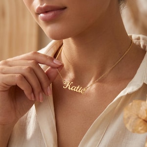 Peut inclure: Un collier doré avec le nom "Katie" en écriture cursive. Le collier est porté autour du cou. La personne porte une chemise blanche boutonnée.