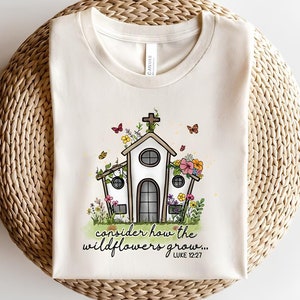 K&ouml;nnte beinhalten: Ein wei&szlig;es T-Shirt mit einem Motiv einer mit Blumen und Schmetterlingen bedeckten Kirche. Der Text "consider how the wildflowers grow... LUKE 12:27" ist unter dem Motiv aufgedruckt.