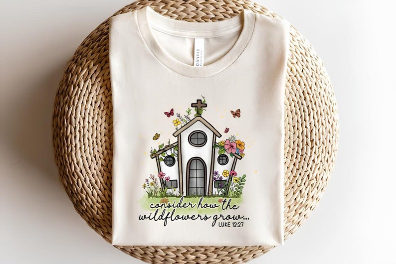 K&ouml;nnte beinhalten: Ein wei&szlig;es T-Shirt mit einem Motiv einer mit Blumen und Schmetterlingen bedeckten Kirche. Der Text "consider how the wildflowers grow... LUKE 12:27" ist unter dem Motiv aufgedruckt.