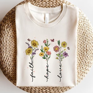 K&ouml;nnte beinhalten: Ein wei&szlig;es T-Shirt mit einem Blumenmuster und den Worten "faith", "hope" und "love", die vertikal geschrieben sind.