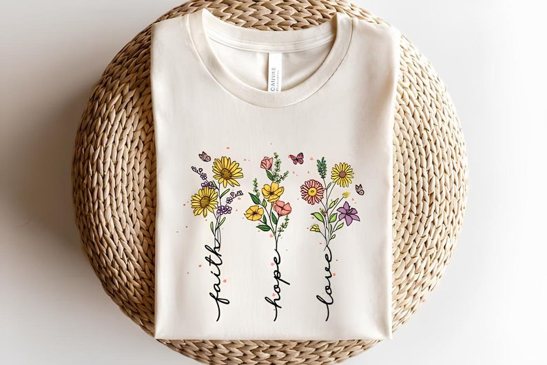 K&ouml;nnte beinhalten: Ein wei&szlig;es T-Shirt mit einem Blumenmuster und den Worten "faith", "hope" und "love", die vertikal geschrieben sind.