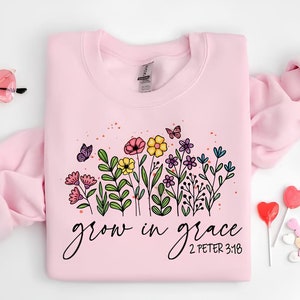 K&ouml;nnte beinhalten: Rosafarbener Pullover mit einem Blumendesign und dem Text "grow in grace 2 Peter 3:18".
