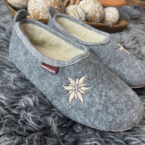 Pode incluir: Chinelos de feltro cinza com uma flor de edelweiss bordada em branco e a palavra "Follkee" no lado. Os chinelos est&atilde;o em um tapete cinza felpudo.