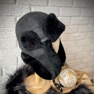 May include: Black leather aviator hat with ear flaps lined in fluffy black fur. The hat is on a wooden display stand.