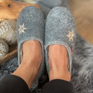 Pode incluir: Um par de pantufas de feltro cinza com flores de edelweiss bordadas. As pantufas est&atilde;o sendo usadas por uma pessoa em um tapete cinza e felpudo.