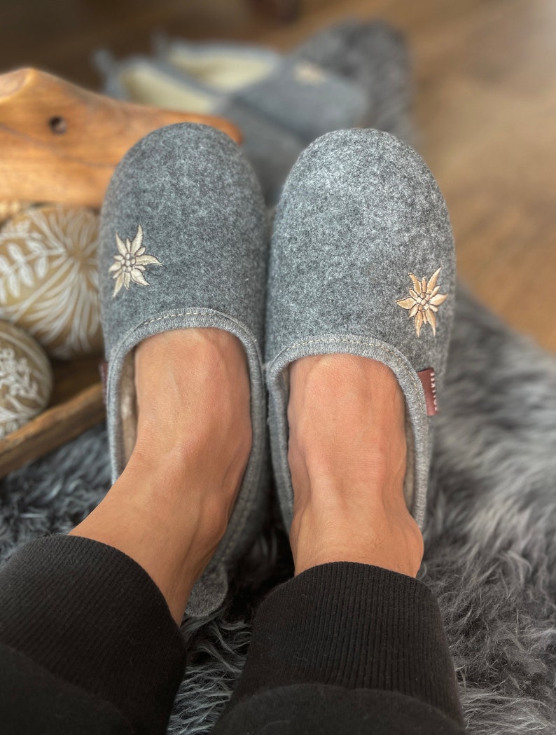 Pode incluir: Um par de pantufas de feltro cinza com flores de edelweiss bordadas. As pantufas est&atilde;o sendo usadas por uma pessoa em um tapete cinza e felpudo.