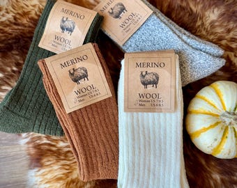May include: A collection of merino wool socks in various colors: olive green, brown, light grey, and cream. Each pair has a ribbed texture and a tag with a sheep illustration and the word "MERINO" and "WOOL".