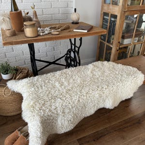 May include: A white sheepskin rug with a fluffy texture, laid out on a wooden floor. The rug is in front of a wooden bench and a table with a white brick wall in the background.