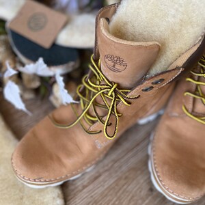 May include: A pair of tan leather boots with yellow laces and a white sole. The boots have a shearling lining and a brown leather logo on the side.