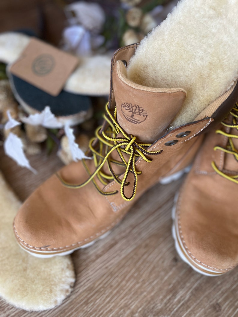 May include: A pair of tan leather boots with yellow laces and a white sole. The boots have a shearling lining and a brown leather logo on the side.