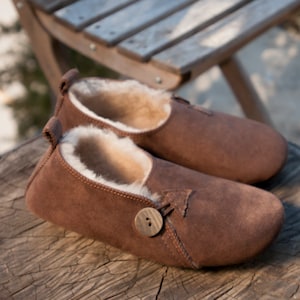 May include: A pair of brown suede slippers with a shearling lining and a wooden button detail.