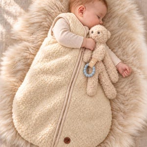 May include: A cream-colored, sleeveless, sherpa-style fleece wearable blanket with a central zipper closure and a small brown circular logo patch at the bottom. A plush, light brown teddy bear toy with a blue silicone teething ring is attached.