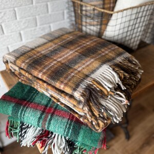 May include: A stack of three plaid blankets in shades of brown, green, and red. The top blanket is brown with a fringe, the middle is green with red stripes, and the bottom blanket is green with red and white stripes. A wire basket with a plaid blanket and a white pillow is in the background.