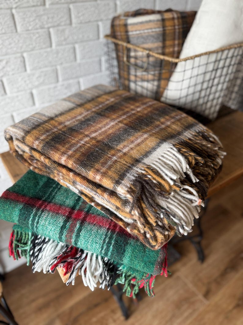 May include: A stack of three plaid blankets in shades of brown, green, and red. The top blanket is brown with a fringe, the middle is green with red stripes, and the bottom blanket is green with red and white stripes. A wire basket with a plaid blanket and a white pillow is in the background.