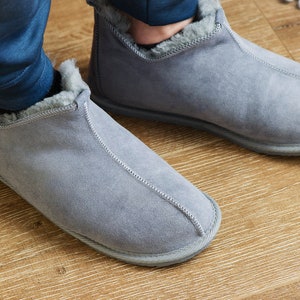 May include: A pair of grey suede slippers with a fluffy lining. The slippers are being worn on a wooden floor.