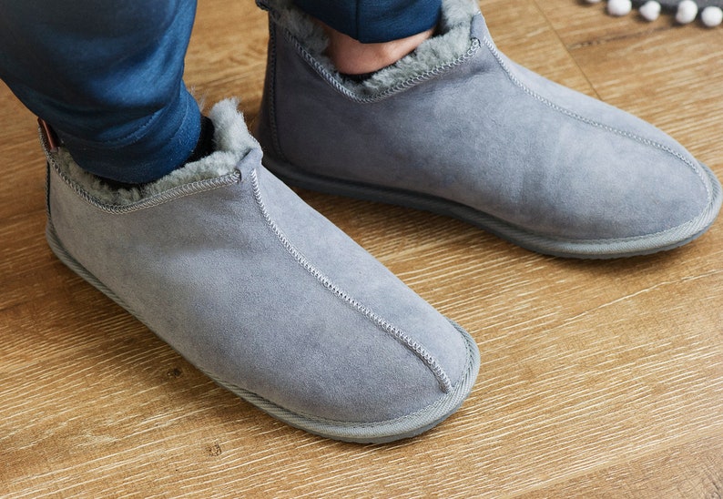 May include: A pair of grey suede slippers with a fluffy lining. The slippers are being worn on a wooden floor.