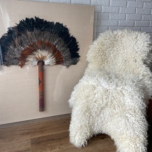 May include: A framed decorative fan made of feathers in shades of black, brown, and white. The fan has a long, patterned handle. A white, curly sheepskin rug is next to the framed fan.