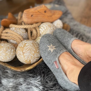 Pode incluir: Chinelos de feltro cinza com um design de flor bordada branca. Os chinelos est&atilde;o em um tapete felpudo com bolas de madeira decorativas.