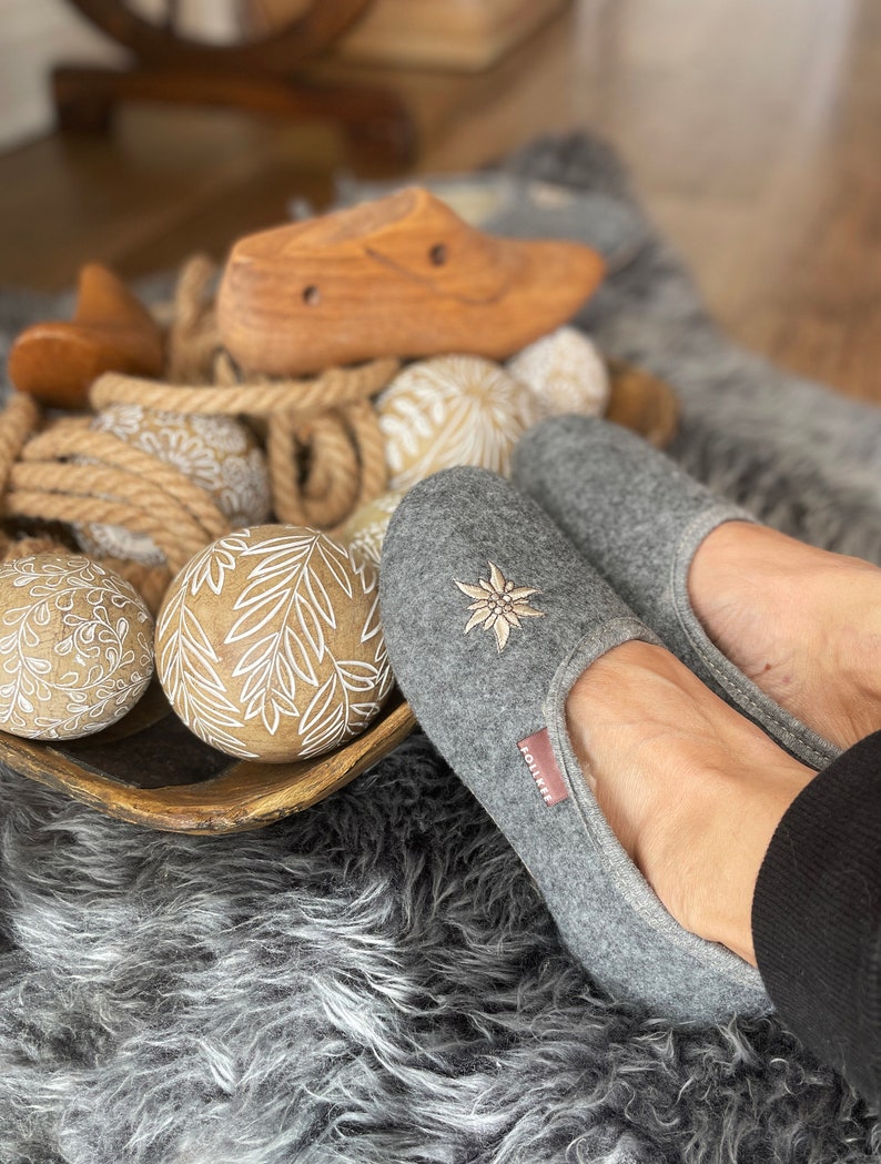 Pode incluir: Chinelos de feltro cinza com um design de flor bordada branca. Os chinelos est&atilde;o em um tapete felpudo com bolas de madeira decorativas.