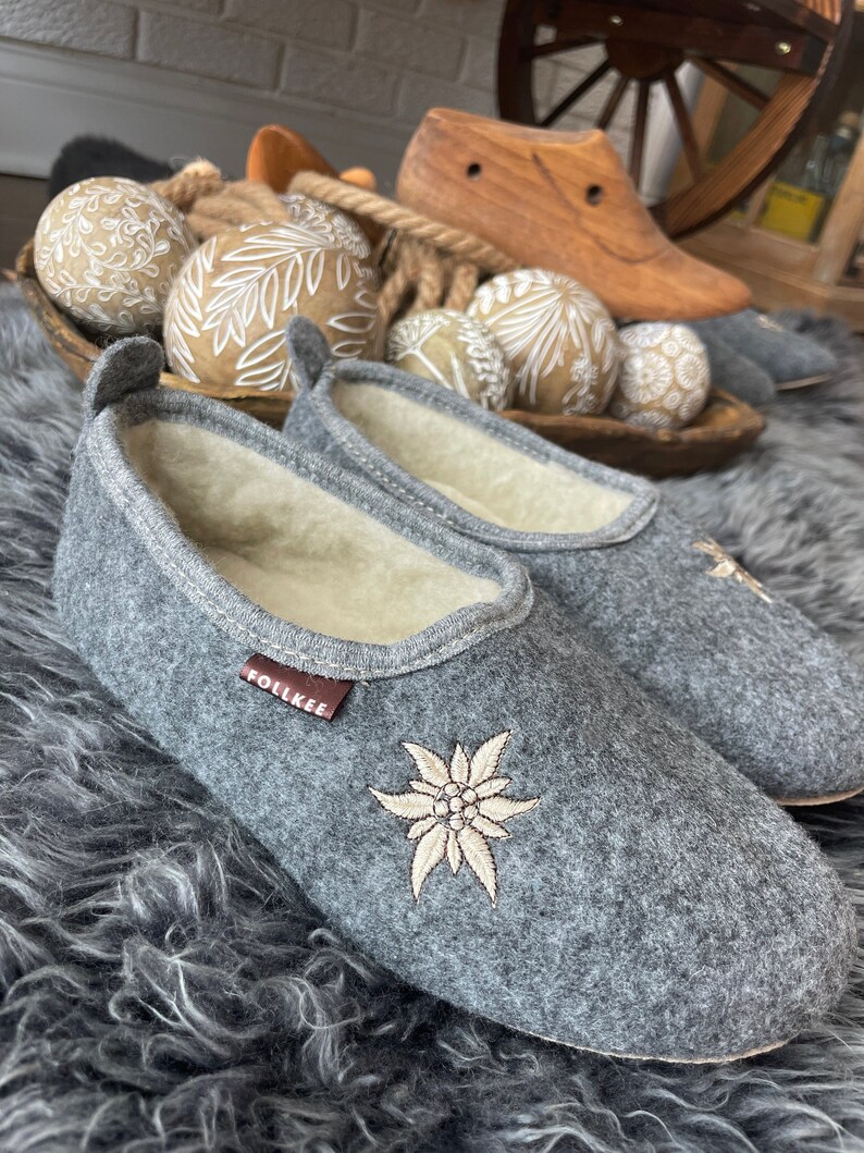 Pode incluir: Chinelos de feltro cinza com uma flor de edelweiss bordada em branco e a palavra "Follkee" no lado. Os chinelos est&atilde;o em um tapete cinza felpudo.