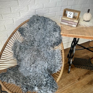 May include: A gray sheepskin rug with a curly texture is placed on a woven chair with a black metal frame. The chair is in front of a white brick wall. A stack of books with a wooden block with the number "2" on it is on a wooden table.