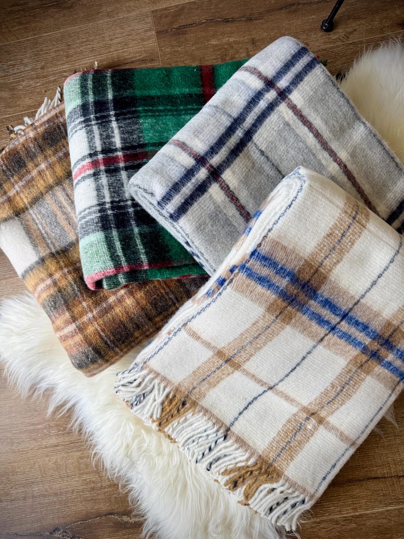 May include: Four folded plaid blankets in shades of brown, green, gray, and white with fringed edges. The blankets are arranged on a white faux fur rug and a wooden surface.