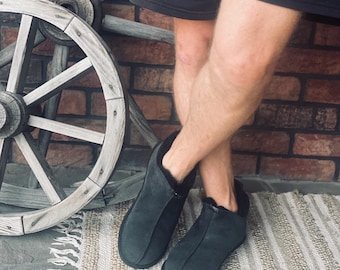 May include: A pair of black suede slippers with a shearling lining. The slippers are worn by a person standing on a rug with a wooden wheel in the background.