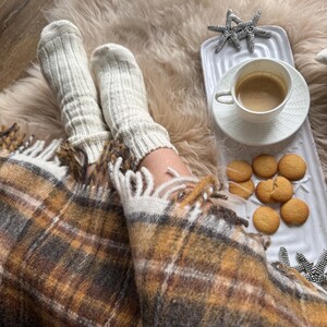 May include: A cozy scene featuring a warm blanket with a plaid pattern in shades of brown, yellow, and white. Cream-colored socks are visible, along with a white tray holding a cup of coffee and cookies. Decorative starfish add a touch of coastal charm.