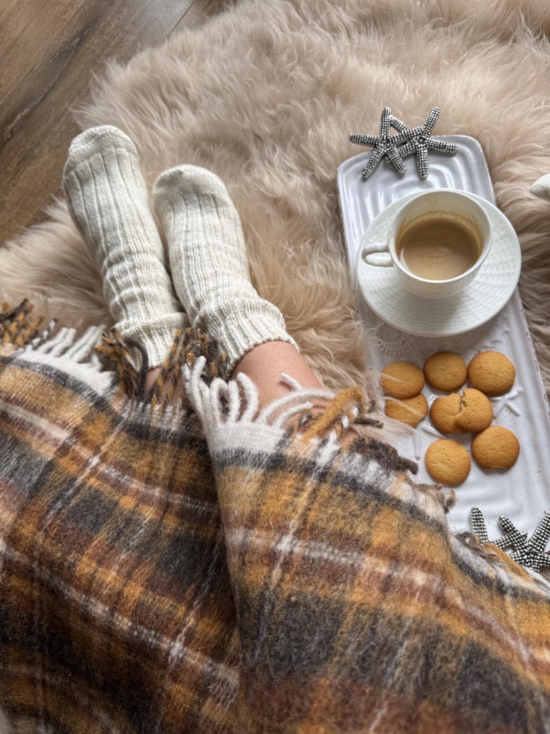 May include: A cozy scene featuring a warm blanket with a plaid pattern in shades of brown, yellow, and white. Cream-colored socks are visible, along with a white tray holding a cup of coffee and cookies. Decorative starfish add a touch of coastal charm.