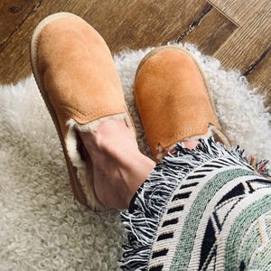 Könnte beinhalten: Ein Paar braune Wildlederslipper mit flauschigem weißen Futter. Die Slipper werden von einer Person getragen, die auf einem weißen, flauschigen Teppich sitzt.